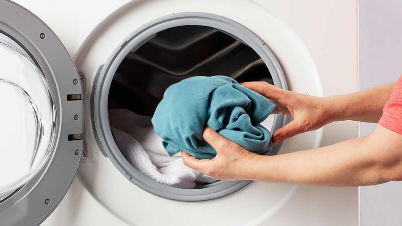 A person loads a blue garment into a front-loading washing machine containing white laundry, showing the care and attention you’d expect from a top laundry service in Toronto.