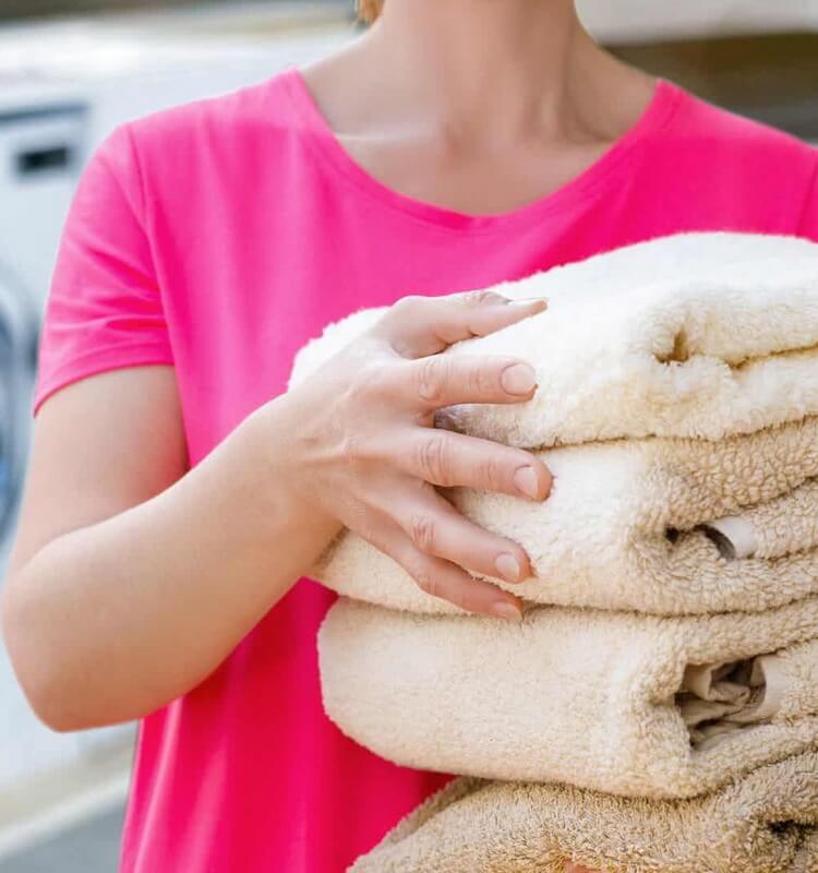 A person holds a stack of folded beige towels while wearing a bright pink shirt, with a washing machine in the background—perfectly capturing the care and attention you can expect from a quality laundry service in Toronto.